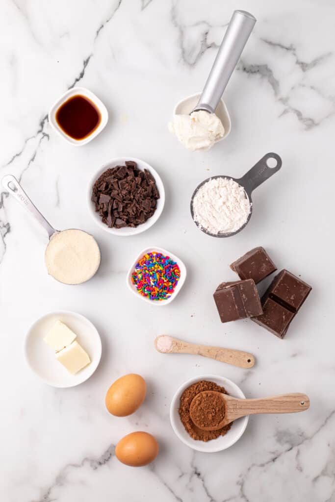 Ingredients for a Choco-Taco recipe arranged on a marble surface, including dark chocolate, cocoa powder, flour, eggs, butter, vanilla, sprinkles, and vanilla ice cream.