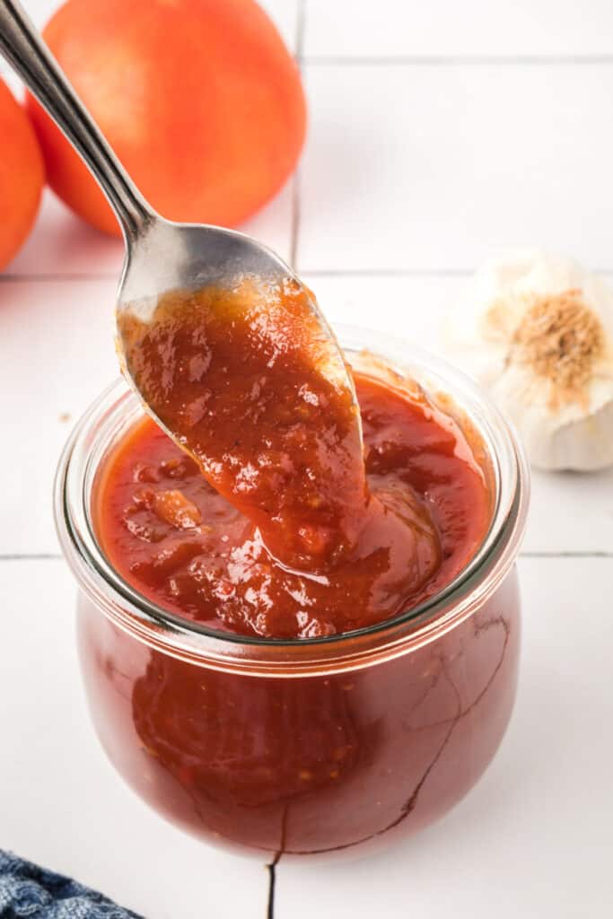 Spoon lifting thick, glossy BBQ sauce from a glass jar