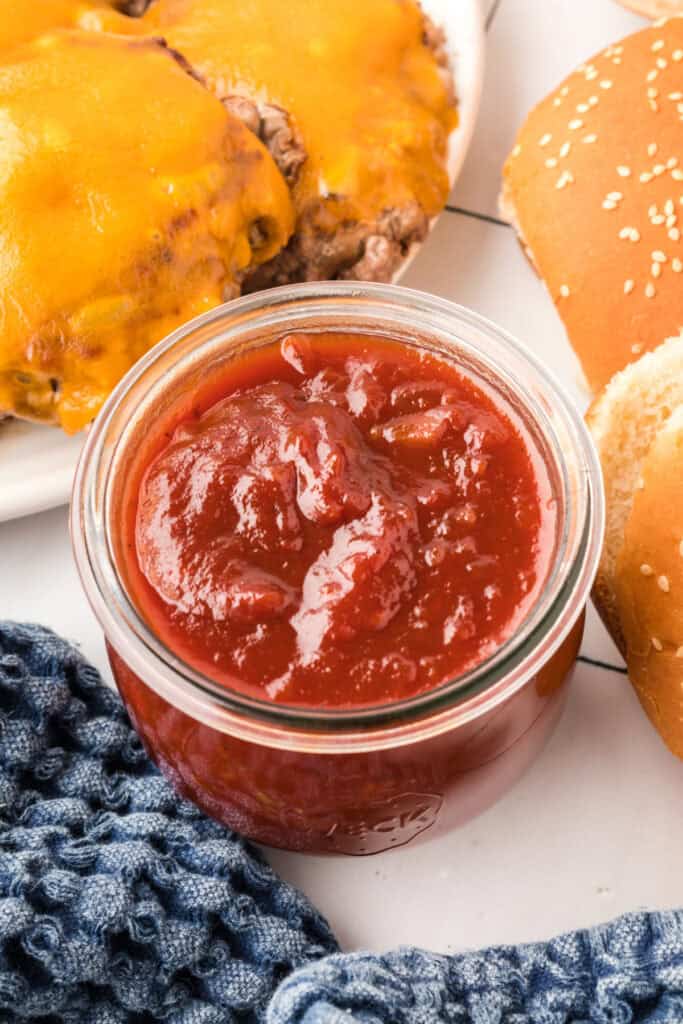 BBQ sauce in a glass jar next to cheddar-topped burgers and sesame bun