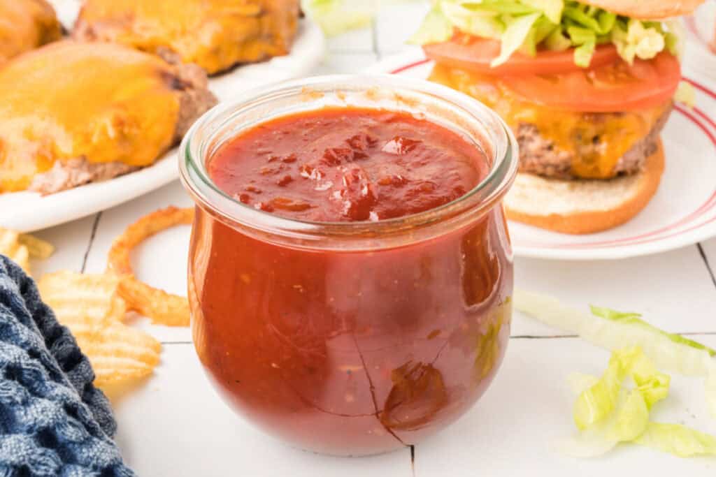 Close-up of BBQ sauce in jar with grilled burgers and lettuce in background