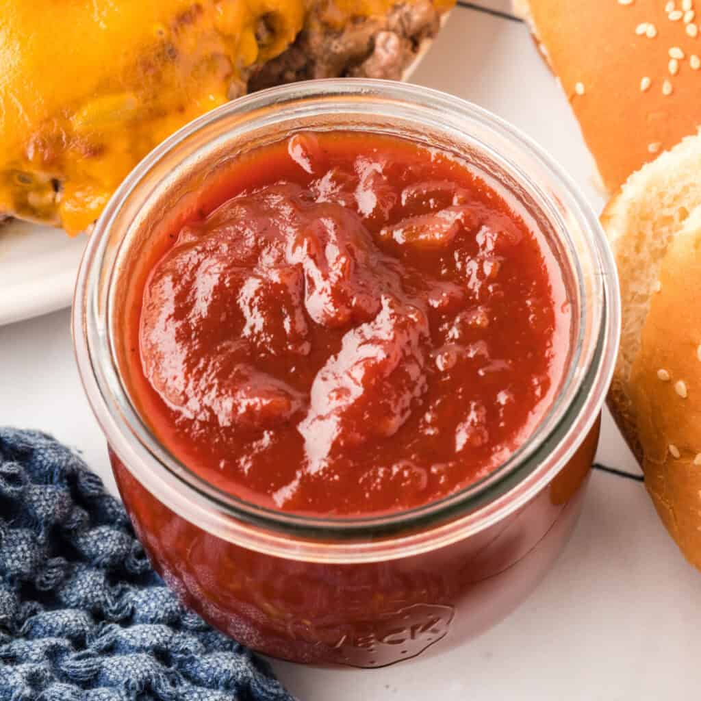 Square crop of jarred BBQ sauce surrounded by burger buns and melted cheese