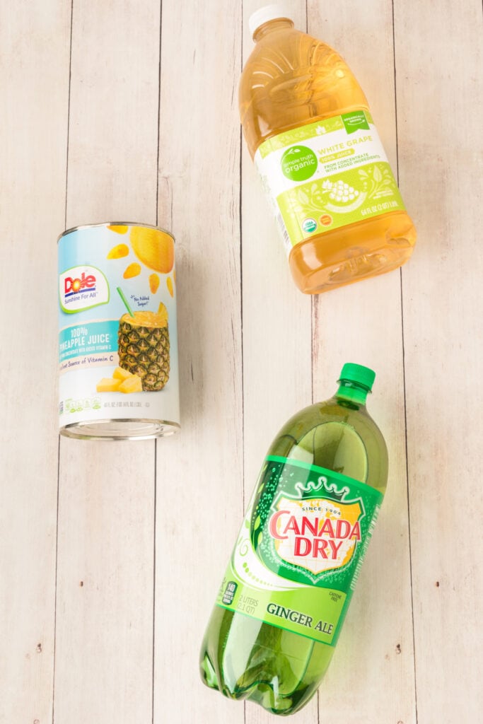 Overhead view of three ingredients for mock champagne punch including pineapple juice, white grape juice, and ginger ale on a light wood surface.