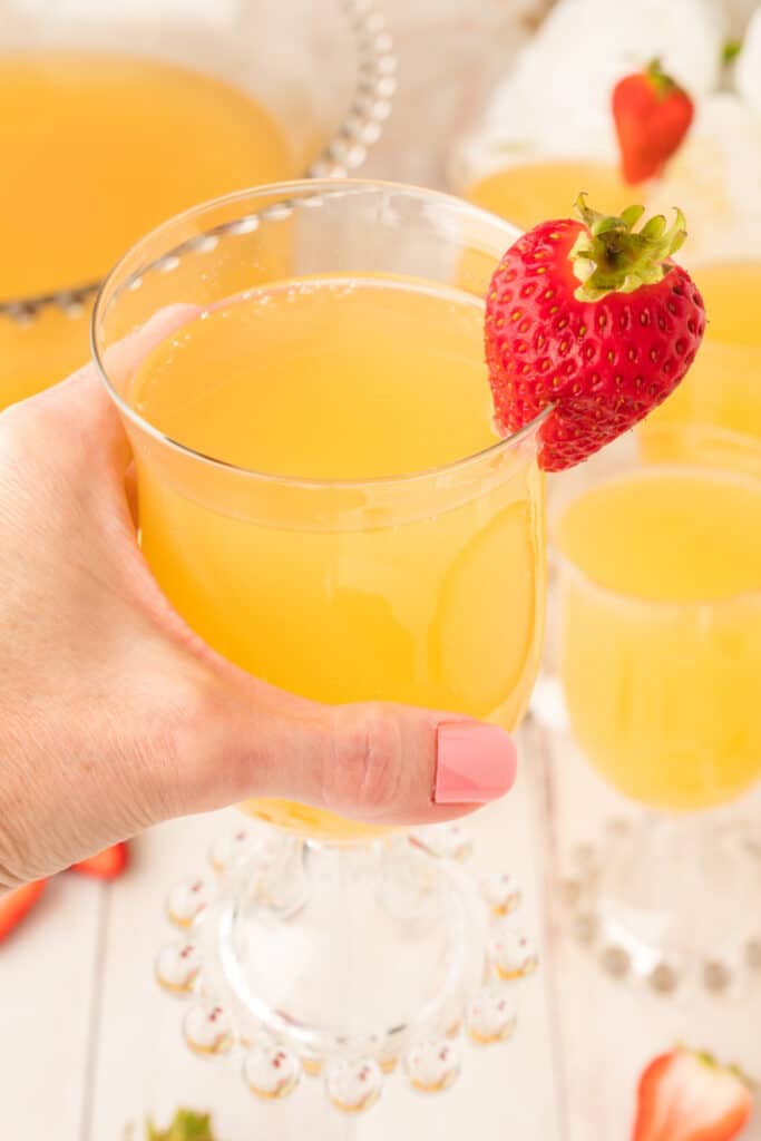 Hand holding a glass of mock champagne punch with a fresh strawberry on the rim and punch bowl in the background.