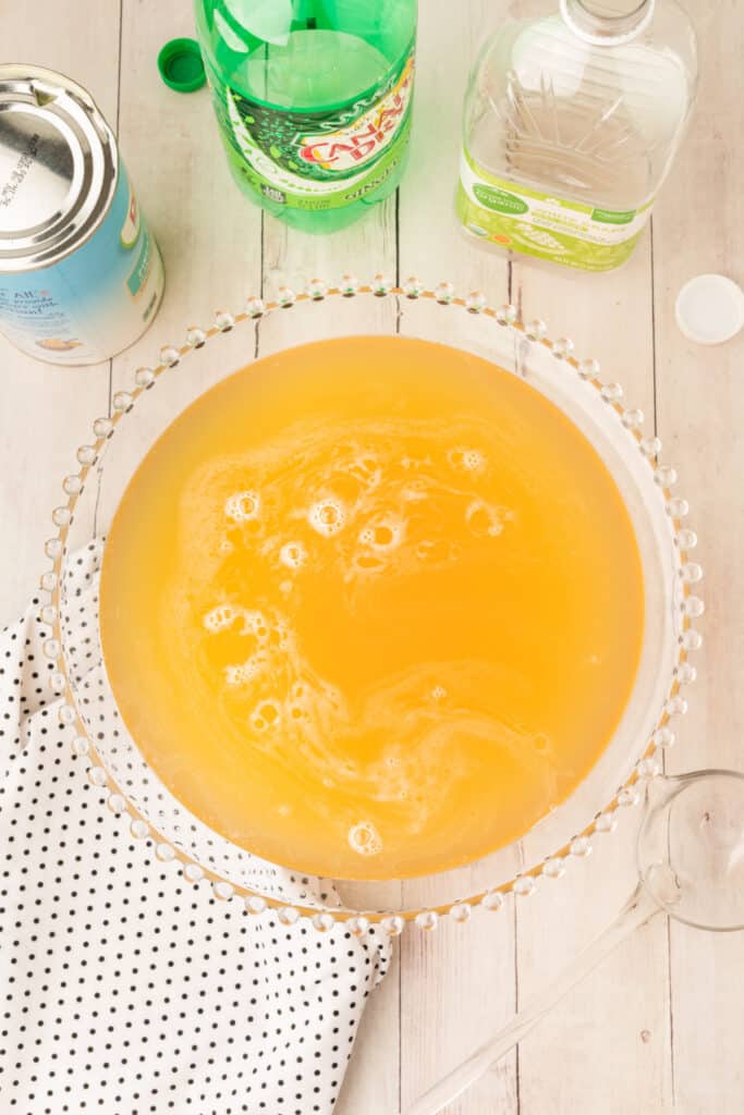 Punch bowl filled with golden mock champagne punch surrounded by empty bottles and cans on a light wood table.