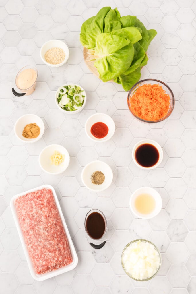 Asian Pork Lettuce Wraps ingredients arranged on a white surface with ground pork, butter lettuce, shredded carrots, sauces, and seasonings