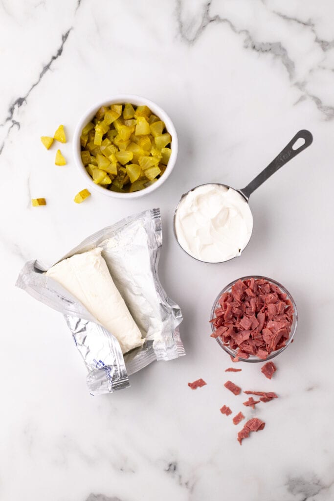 Overhead view of four main ingredients: chopped pickles, sour cream, cream cheese, and dried beef on a marble surface