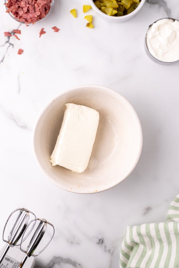 Cream cheese block in a white mixing bowl next to ingredients, hand mixer in view