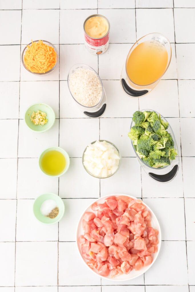Ingredients for Cheesy Chicken Broccoli Rice Skillet arranged on a white tile countertop including chicken, broccoli, rice, broth, cheese, onion, garlic, and cream of chicken soup