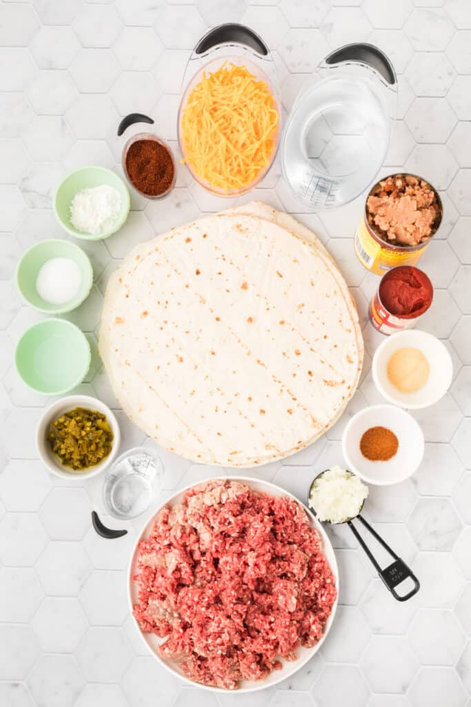 Ingredients for Chili Cheese Burrito arranged on a white countertop with ground beef, flour tortillas, cheddar cheese, refried beans, tomato paste, seasonings, onion, jalapeños, cornstarch, and water.