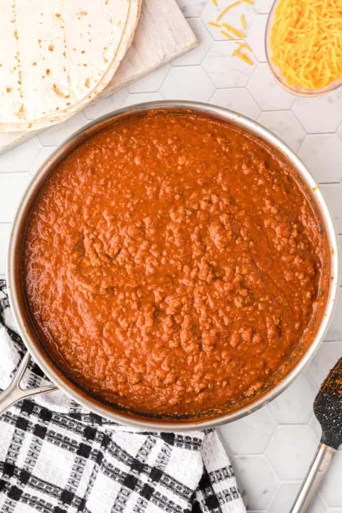 Finished Chili Cheese Burrito filling in a large skillet with flour tortillas and shredded cheddar cheese nearby.