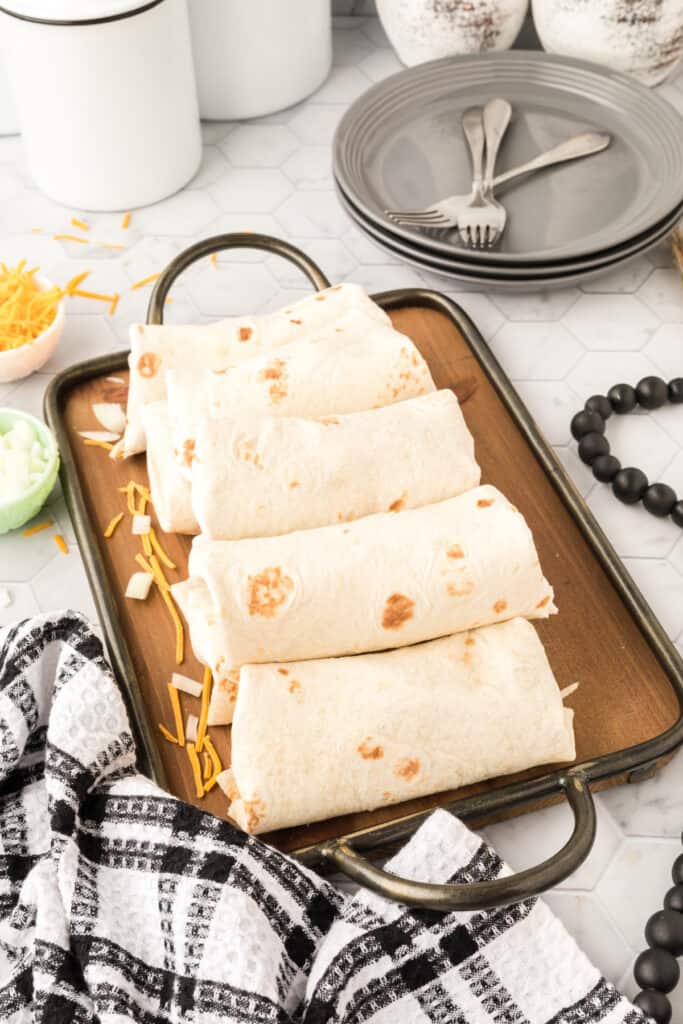 Rolled Chili Cheese Burritos arranged on a serving tray with shredded cheddar cheese and diced onion nearby.