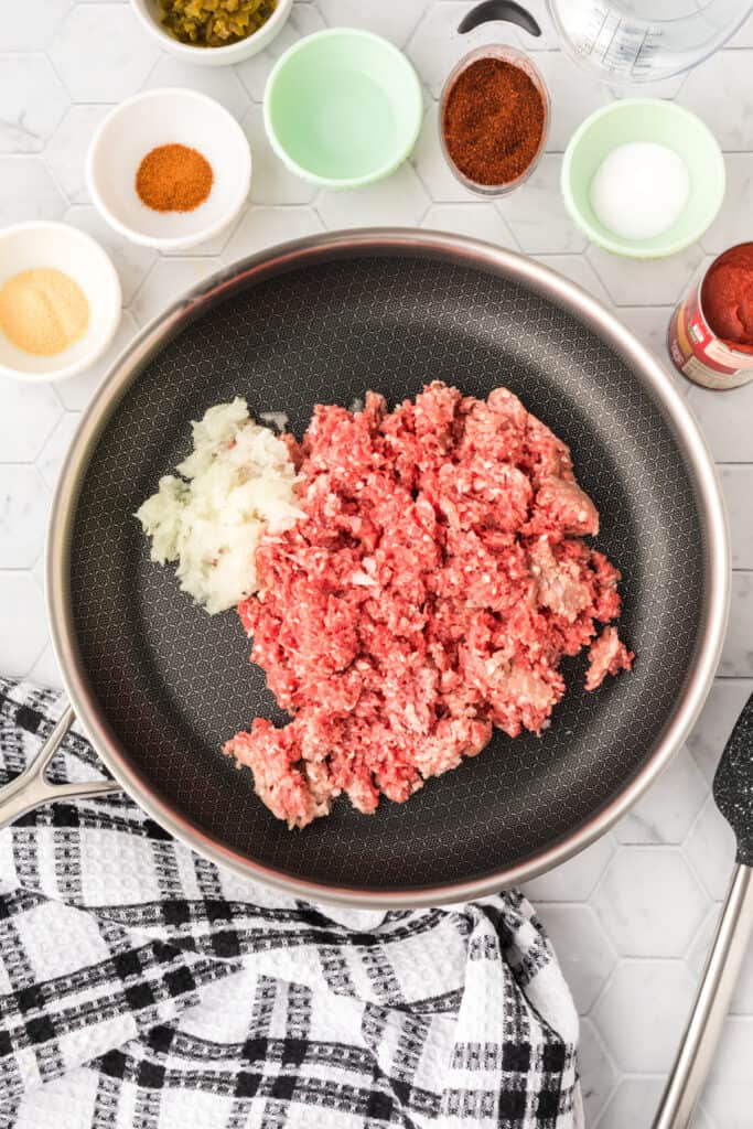 Raw ground beef and chopped onion in a large skillet with measured Chili Cheese Burrito ingredients surrounding the pan.