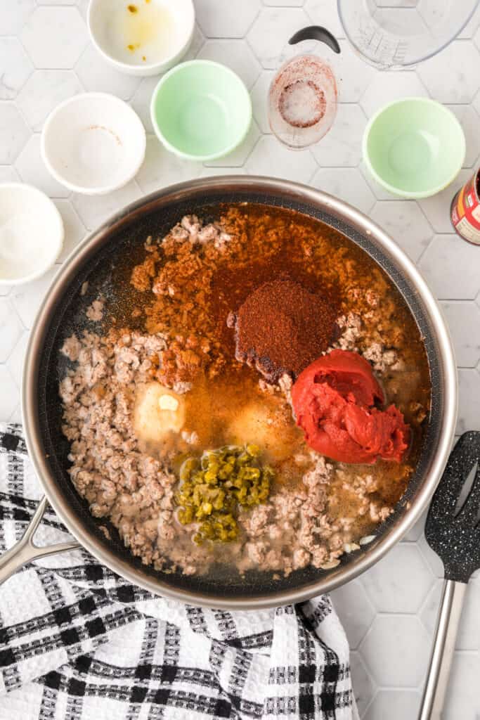 Ground beef in a skillet topped with tomato paste, chili powder, jalapeños, seasonings, vinegar, and water before simmering.