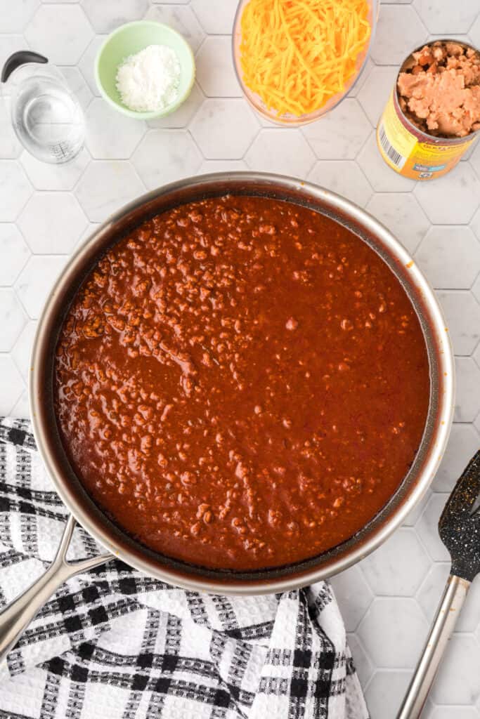 Chili Cheese Burrito filling simmering in a large skillet with cheddar cheese, refried beans, cornstarch, and water nearby.