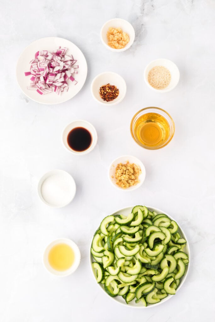 sliced cucumbers, chopped red onion, soy sauce, rice vinegar, sesame oil, garlic, ginger, sesame seeds, sugar, and red pepper flakes arranged on a white surface