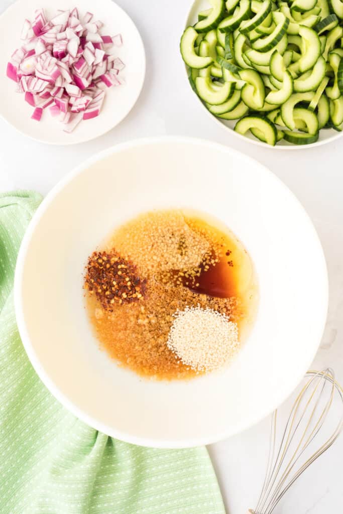 dressing ingredients including soy sauce, sesame oil, sesame seeds, garlic, ginger, and red pepper flakes in a white mixing bowl with sliced cucumbers and chopped onion nearby