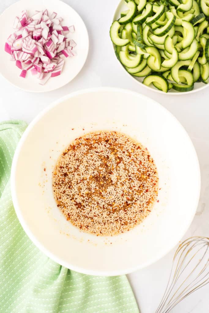 whisked dressing with sesame seeds and spices in a white bowl with sliced cucumbers and chopped red onion beside it