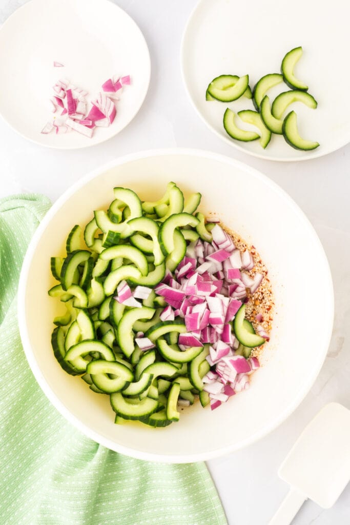 sliced cucumbers and chopped red onion added to a bowl with prepared dressing