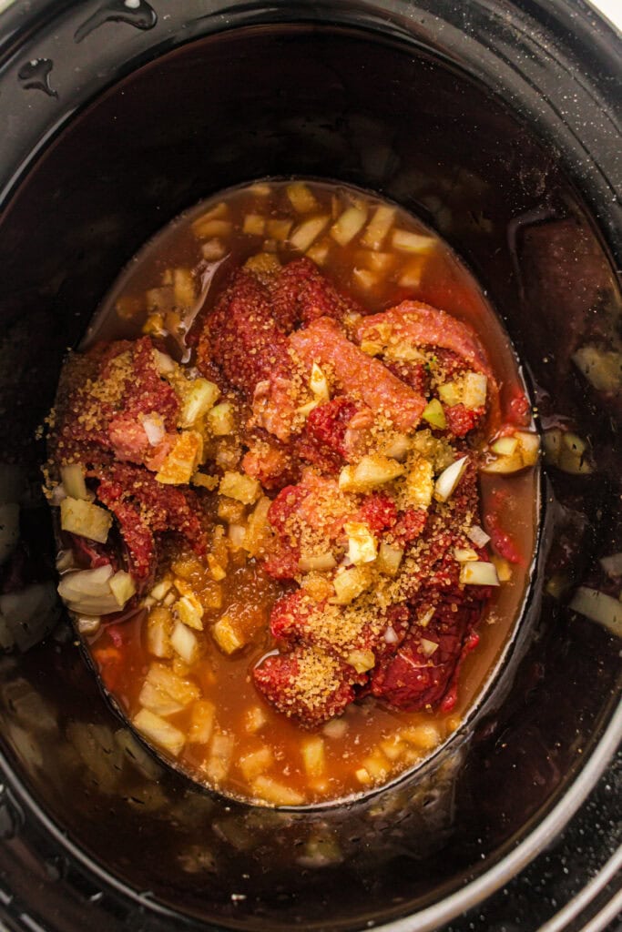 Raw sliced beef with diced onion, garlic, brown sugar, and sauce inside a slow cooker before cooking