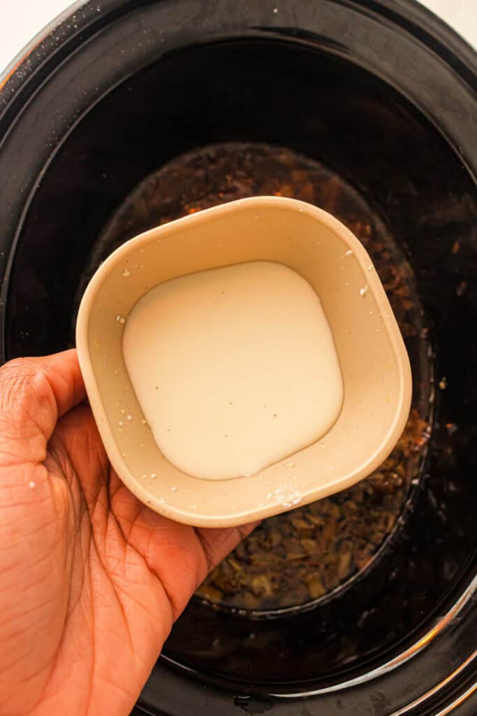 Hand holding a small bowl of cornstarch slurry over cooked beef mixture in a slow cooker