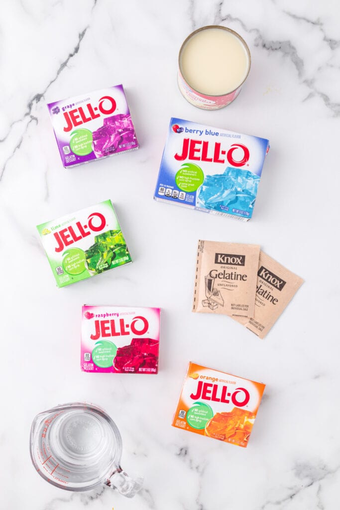 Ingredients for Broken Glass Jello recipe including colorful Jello boxes, sweetened condensed milk, Knox gelatin, and measuring cup on marble background