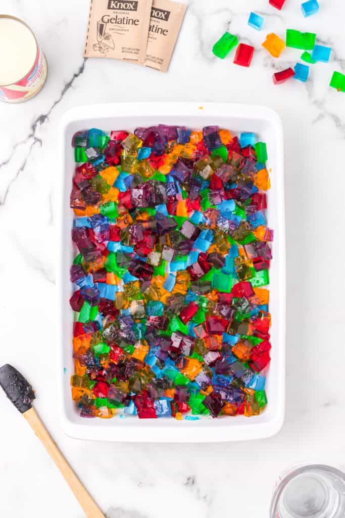 Mixed colorful gelatin cubes in baking dish for Broken Glass Jello recipe with sweetened condensed milk nearby