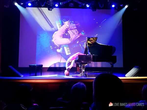 A man laying upside down on a piano while playing it on the stage of a cruise ship.