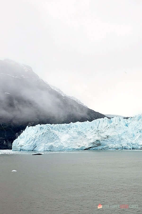 An iceberg sits in a body of water with mountains in the background.