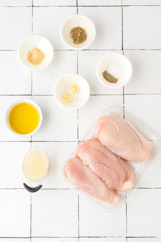 Overhead view of raw chicken breasts with small bowls of marinade ingredients on white tile background