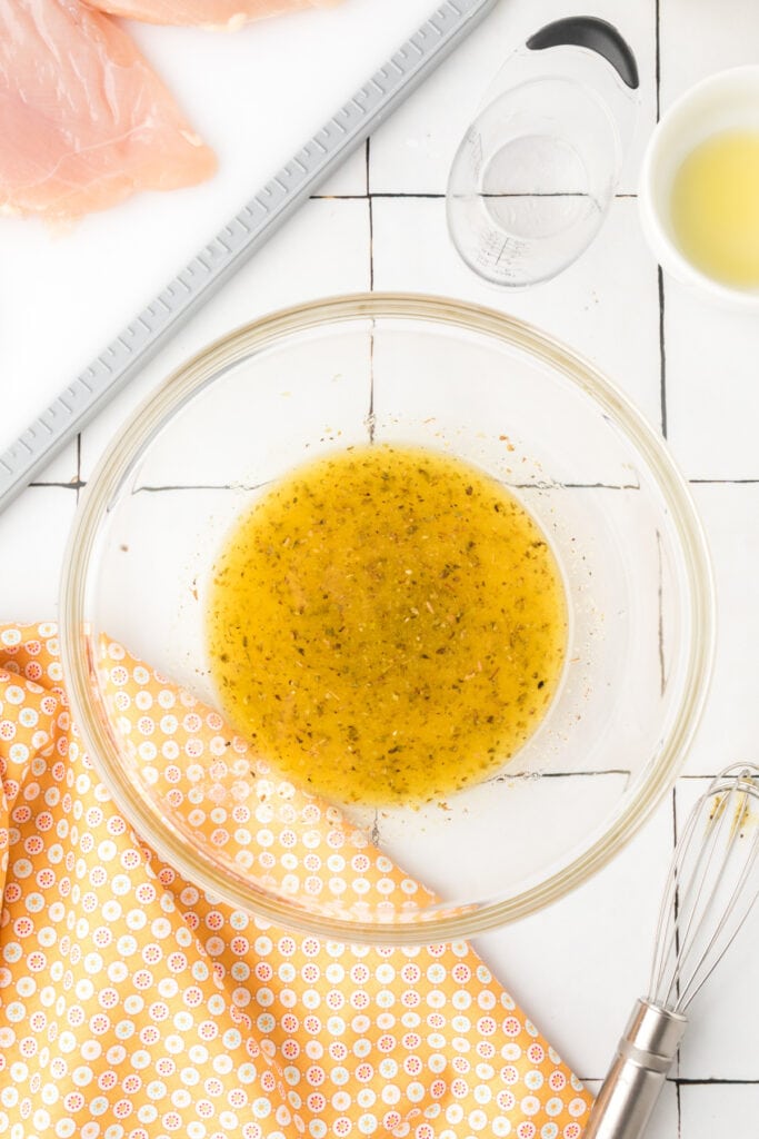 Fully mixed grilled chicken marinade in a clear glass bowl on a white tile background