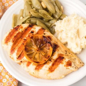 Square-cropped image of grilled chicken breast with grill marks and lemon slice, served with mashed potatoes and green beans