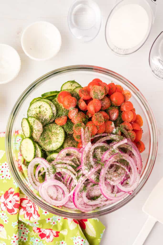 Salad ingredients in a glass bowl topped with fresh dill, salt, and celery seed