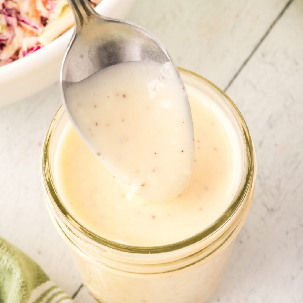 Overhead view of a spoon lifting creamy coleslaw dressing from a jar with a green towel nearby.