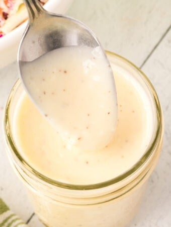 Overhead view of a spoon lifting creamy coleslaw dressing from a jar with a green towel nearby.