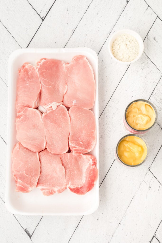 Package of raw pork chops on a white wood surface next to bowls of dry Ranch mix and opened cans of cream of chicken soup.