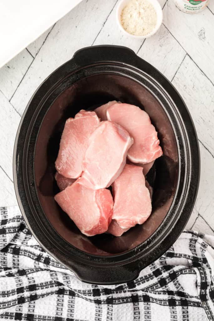 Uncooked pork chops placed inside a slow cooker with dry Ranch seasoning and soup ingredients nearby.