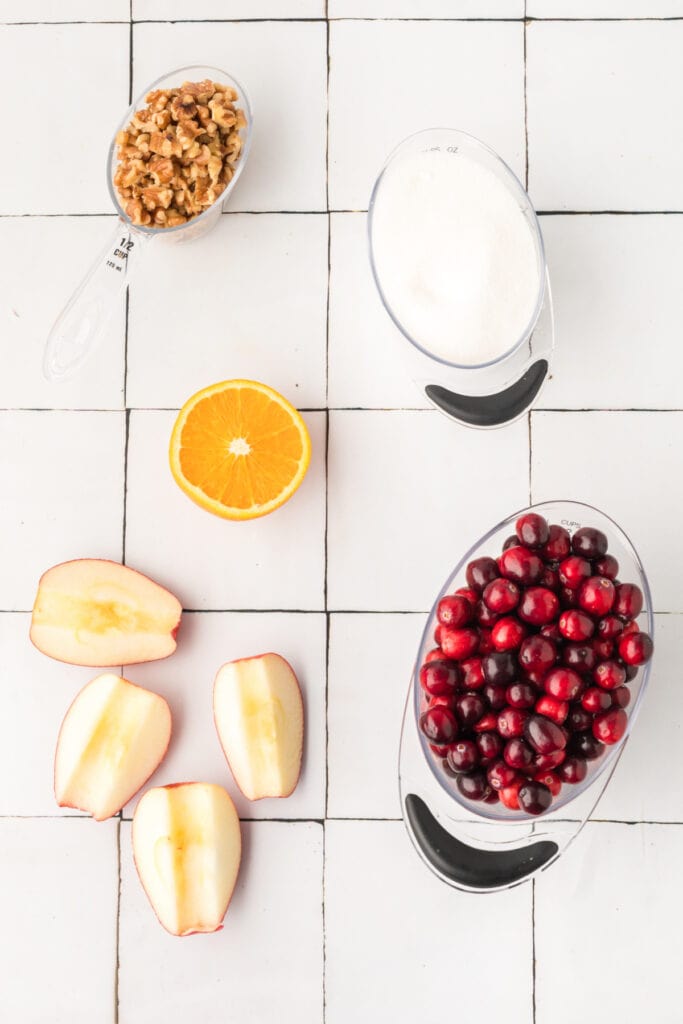 Overhead view of fresh ingredients for cranberry relish including chopped walnuts, sugar, half an orange, apple quarters, and cranberries arranged on a white tiled surface.