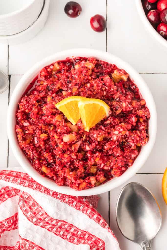 Bright cranberry relish in a white serving bowl garnished with orange slices, with a red-striped towel and silver spoon beside it.