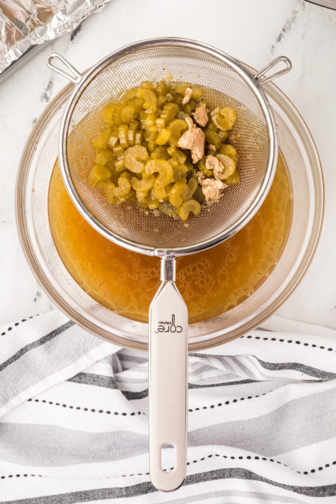 Strainer with cooked celery and bits of turkey over a glass bowl of broth for gravy preparation.