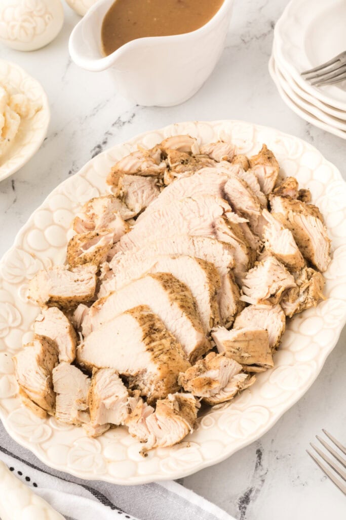 Sliced Instant Pot turkey breast arranged on a white platter with a gravy boat, mashed potatoes, and stacked plates nearby.
