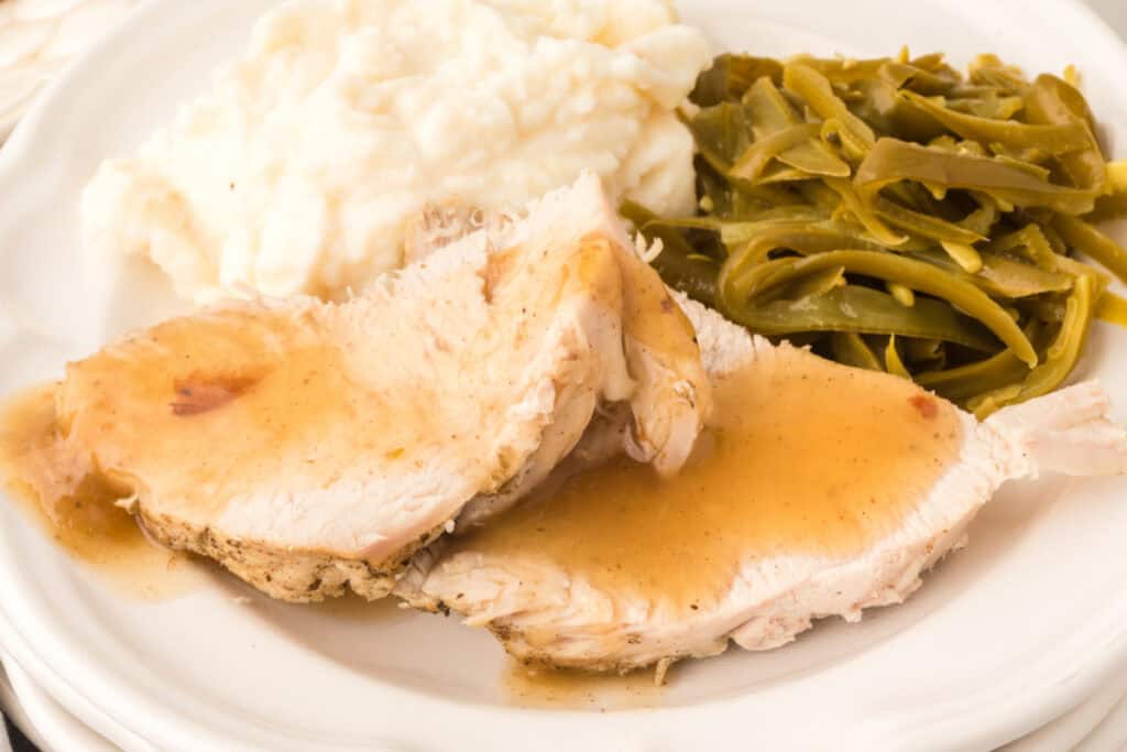 Close-up of sliced turkey breast topped with gravy, served with mashed potatoes and green beans on a white dinner plate.