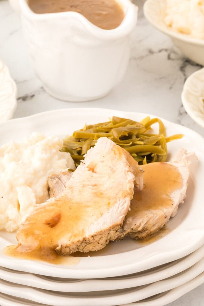 Plated serving of turkey breast with gravy, mashed potatoes, and green beans, with a gravy boat visible in the background.