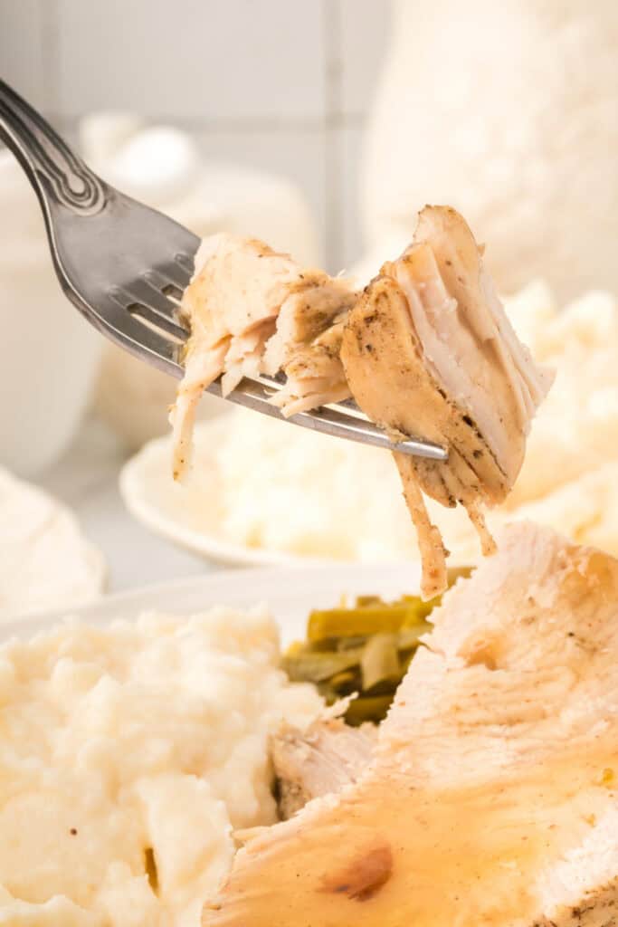Fork lifting a bite of tender turkey breast with mashed potatoes and green beans blurred in the background.
