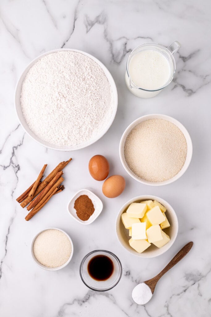 overhead view of Amish Cinnamon Bread ingredients on a marble surface including flour, buttermilk, sugar, eggs, butter, vanilla, cinnamon sticks, ground cinnamon, and baking soda