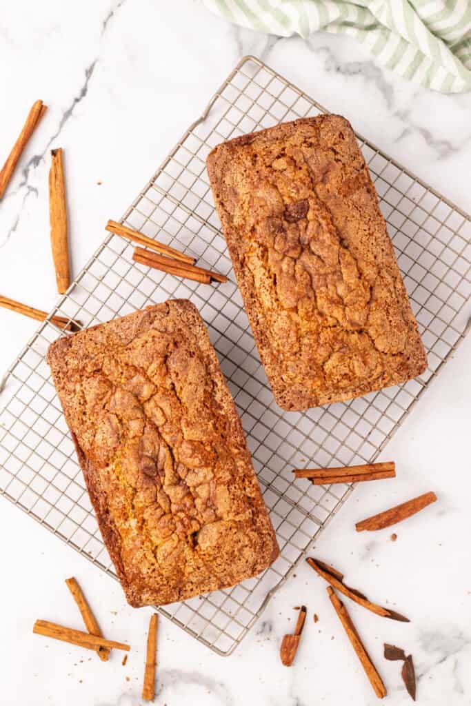 two baked Amish Cinnamon Bread loaves cooling on a wire rack with cinnamon sticks scattered around