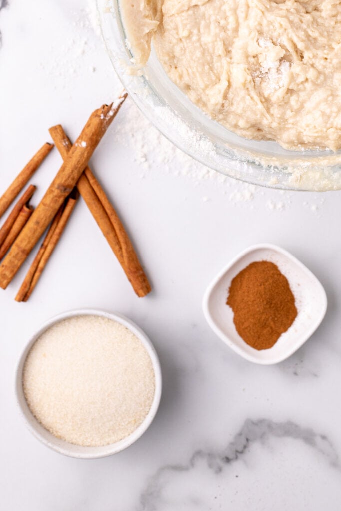 overhead view of the cinnamon-sugar topping ingredients - granulated sugar, ground cinnamon, and cinnamon sticks - next to the prepared bread batter