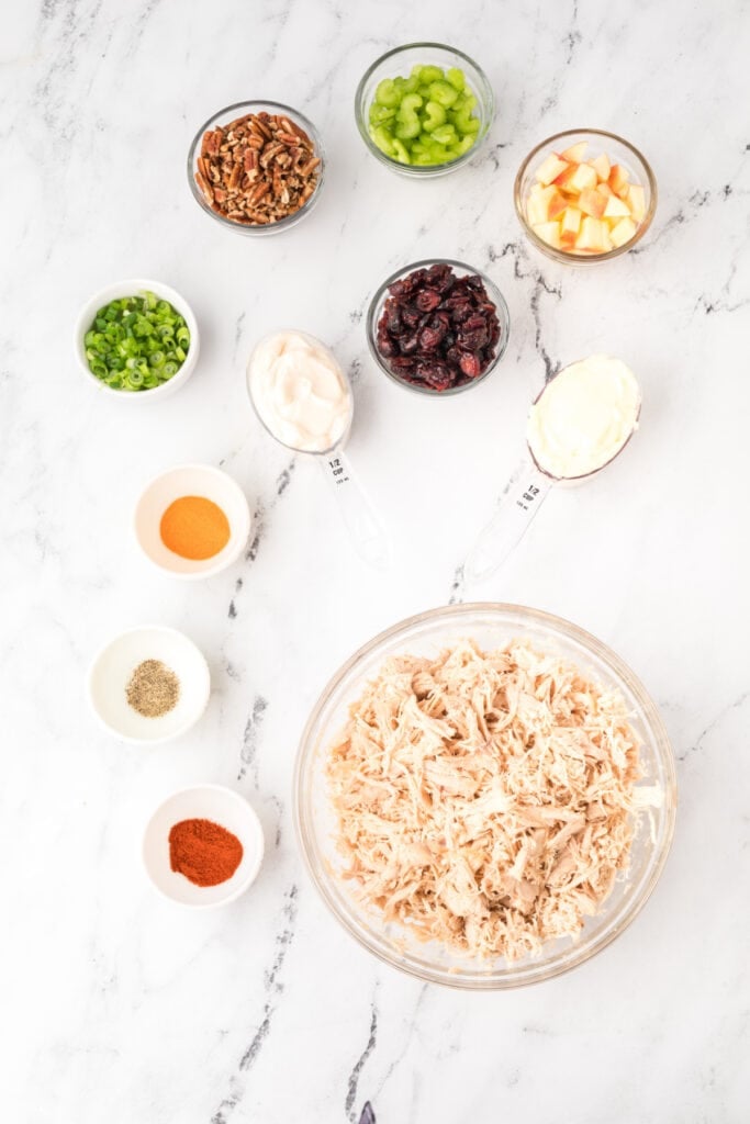 ingredients for chicken salad with Miracle Whip including shredded chicken, celery, pecans, dried cranberries, green onions, apple, and dressing on a white surface