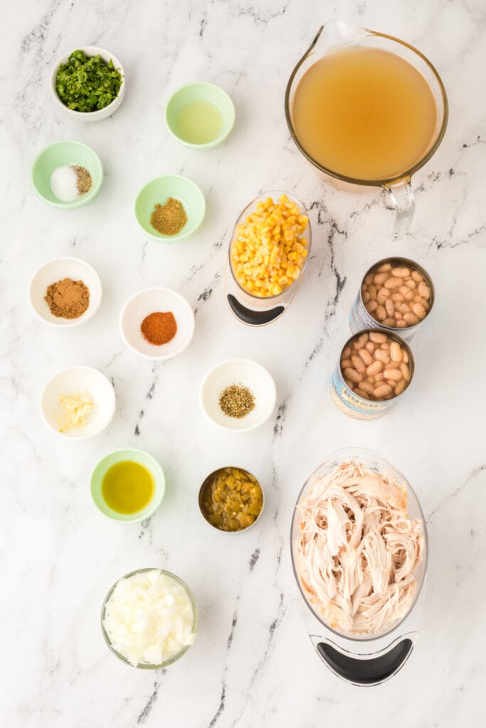 Overhead shot of White Chicken Chili ingredients in small bowls and measuring cups on a marble surface, including corn, beans, broth, and spices.