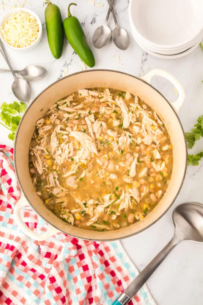 Overhead view of White Chicken Chili with shredded chicken, white beans, and corn in a cream-colored Dutch oven surrounded by jalapeños, shredded cheese, spoons, and bowls