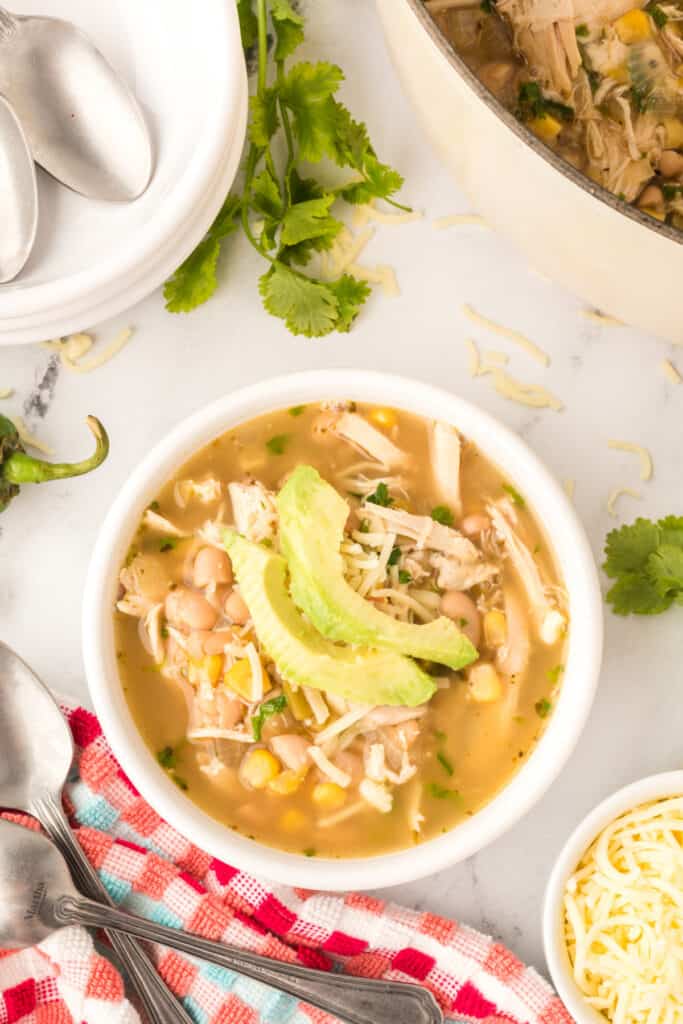 Overhead bowl of White Chicken Chili topped with sliced avocado, shredded cheese, and cilantro with a colorful towel and spoons beside it.
